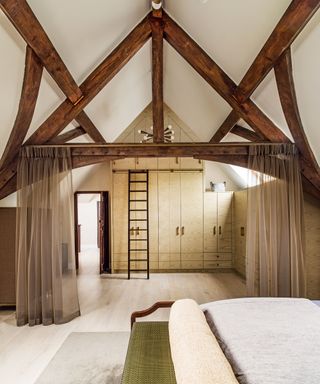 Master bedroom with a wall of storage and sliding ladder to reach the top level, wardrobes and exposed beams under a vaulted ceiling, and net curtains drawn back.