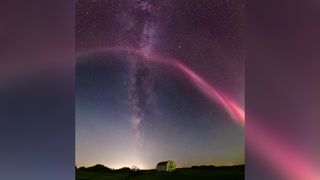 a long red stream of light arches over the night sky, in the background is the milky way appearing as a thick band of stars running from the top of the image down to the horizon.