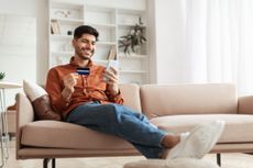Smiling young man holding credit card in hand and using cell phone at home