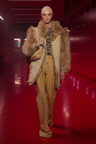 A model walking in the F/W 25 Valentino show during Paris Fashion Week wearing a tan fur coat with a patterned blouse and mustard trousers.