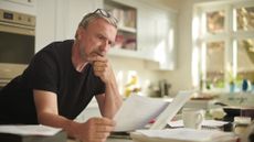 A man in his early 60s leans on his kitchen island, reading a bill or financial statement and looking concerned. 
