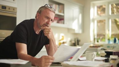A man in his early 60s leans on his kitchen island, reading a bill or financial statement and looking concerned. 