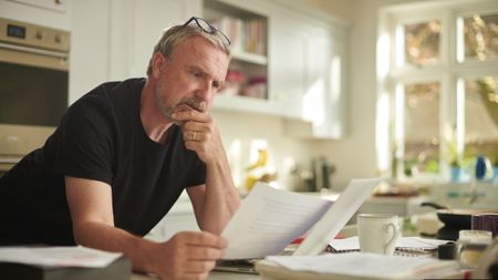 A man in his early 60s leans on his kitchen island, reading a bill or financial statement and looking concerned. 