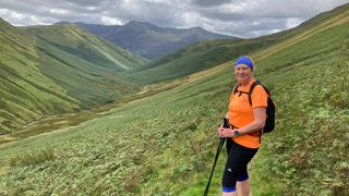 A woman trail running gear and holding running poles stands in front of a scenic mountain view