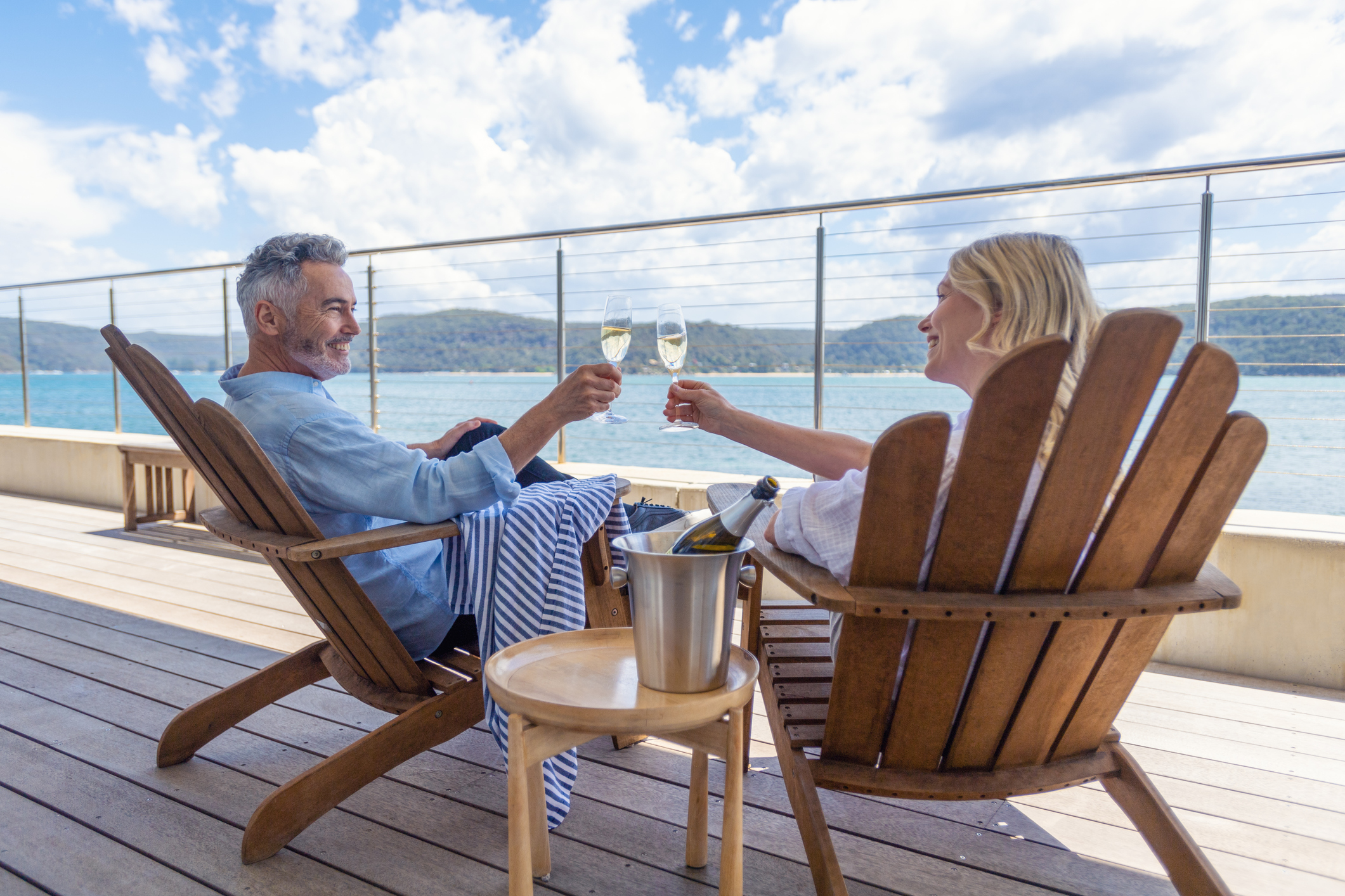 Mature couple drinking champagne out on the deck. They are happy and smiling sitting in deck chairs. The sea is in the background