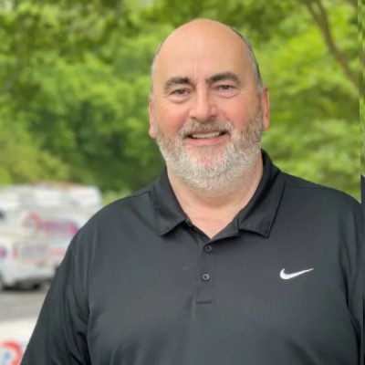 Gary is a white man with a light beard pictured wearing a black Nike polo shirt. He appears to be standing in the company car park with logos visible (but blurred) on white vans in the background