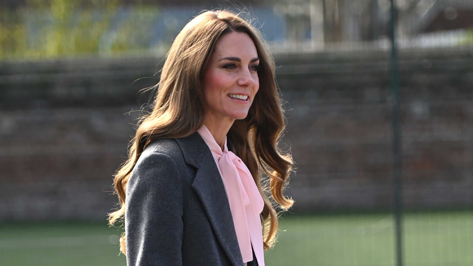 Kate Middleton walks outside wearing a grey coat with a pink blouse, grey trousers and grey high heels.