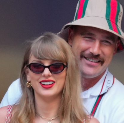 Taylor Swift wearing sunglasses and smiling while Travis Kelce hugs her from behind wearing a bucket hat and white Gucci polo