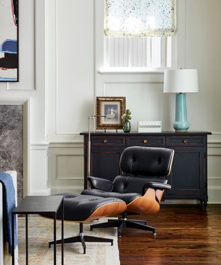 white living room with paneling, Eames chair, black dresser, hardwood floor, rug, aqua table lamp, blind, artwork