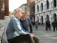 Mature older gay male couple living abroad, sitting on stone steps of a historic building in European square on a sunny afternoon, looking upwards toward the top of buildings or sky.