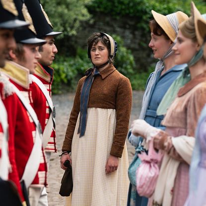 A Regency-era woman (Ella Bruccoleri as Mary Bennet) stands alone while framed by a row of red-coated soldiers and another row of Regency women, in BritBox's 'The Other Bennet Sister.'