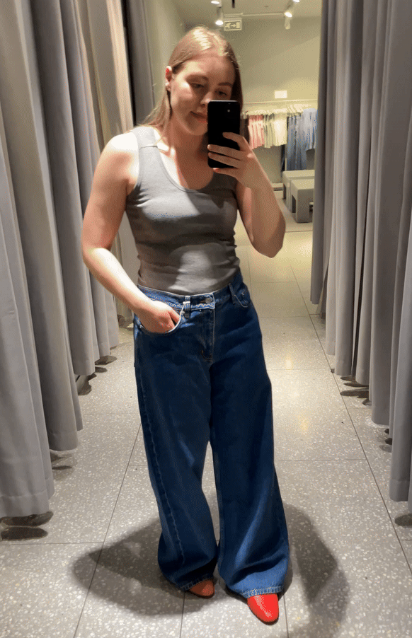 Woman in mirror wears grey vest, blue jeans and red shoes