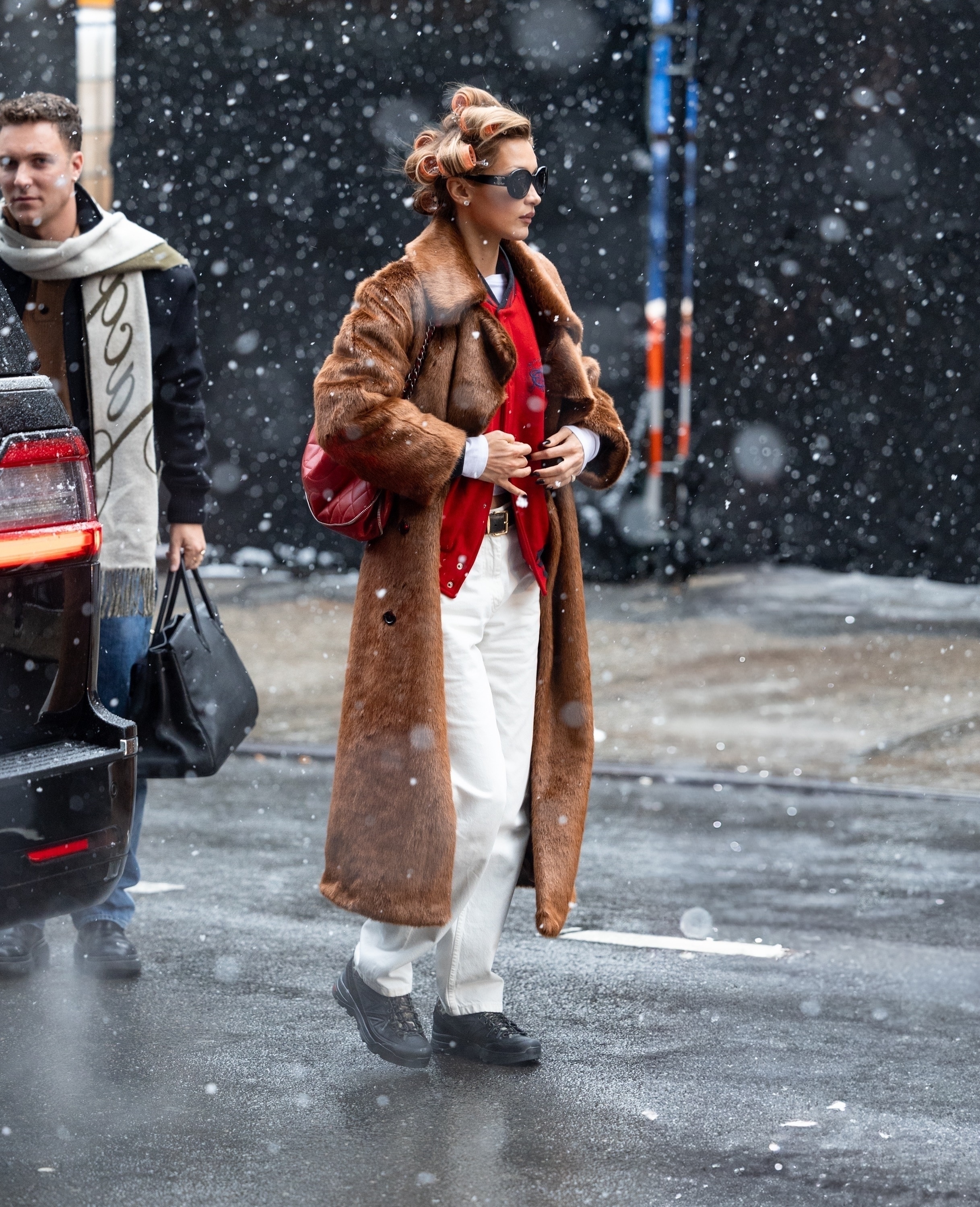 Bella Hadid wears a fur coat, red sweater, white jeans, and black sneakers.