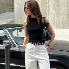 fashion influencer Débora Rosa poses in front of a vintage car wearing sunglasses, a black tank top, a black shoulder bag, Khaite studded belt, and white pants