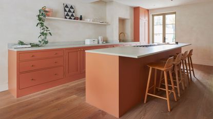 A kitchen in a peach/orange color with a white colored polished concrete worktop. There is also an island with four bar stools beside it. On one side of the kitchen there is also an open shelf with a plant and art work on top
