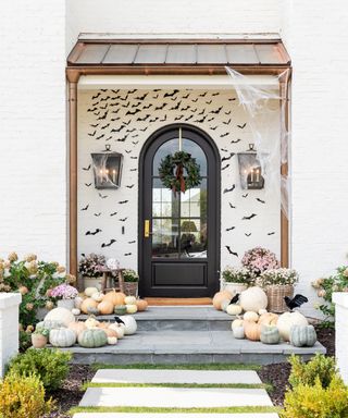 Halloween door decor ideas with pale pumpkins on the floor, black bats stuck to the houses's white walls, and a fake web hanging from the porch roof