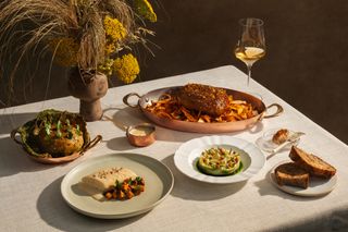 Close up view of food including bread, fish and meat and a glass of wine on a table at Claudine, Singapore. The table features a light grey table cloth and a plant in a brown pot. The background is dark brown