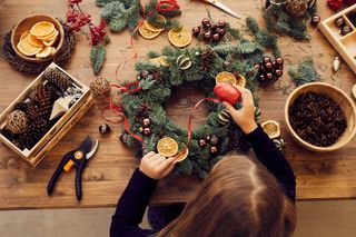 A image from above of a girl making a Christmas wreath at a desk with fresh pine, pine cones, dried oranges, and ribbon.