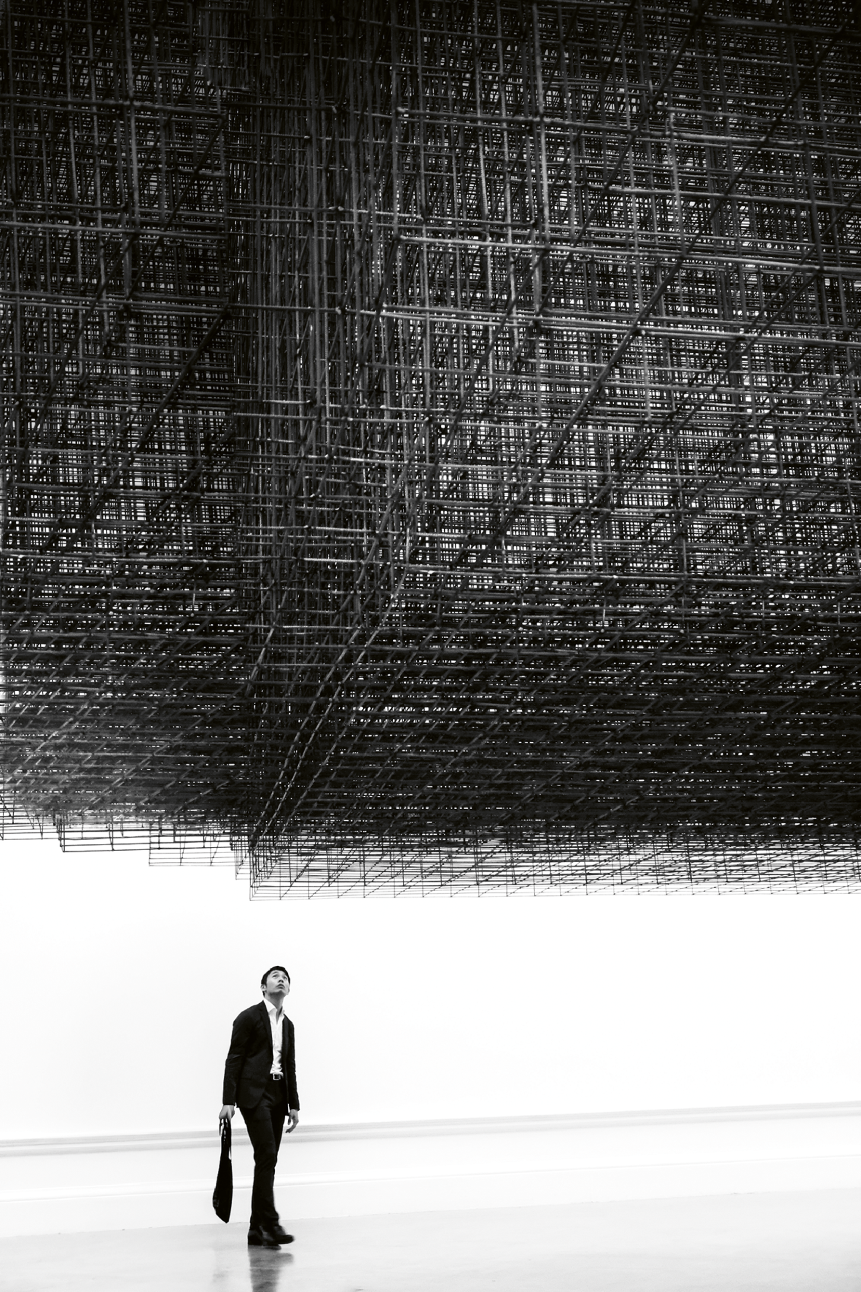 A man in a black suit carrying a bag looks up in a white gallery space at a huge dark lattice of metal rods suspended overhead like a cloud.