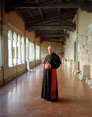 Photographic portrait of Cardinal in black and red robes pictured in cloisters.