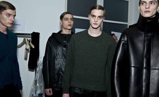 Four male models wearing looks from Jil Sander's collection. They are wearing a black jacket, a thick black panelled coat, a green jumper and a blue piece with a crew neck and front zip