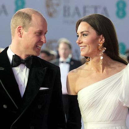 Prince William wears a tuxedo while his wife Kate Middleton wears a one-shoulder white gown with long dangly gold earrings and long black gloves