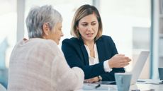 An older woman listens in an office while a financial adviser talks and points at her laptop screen. 