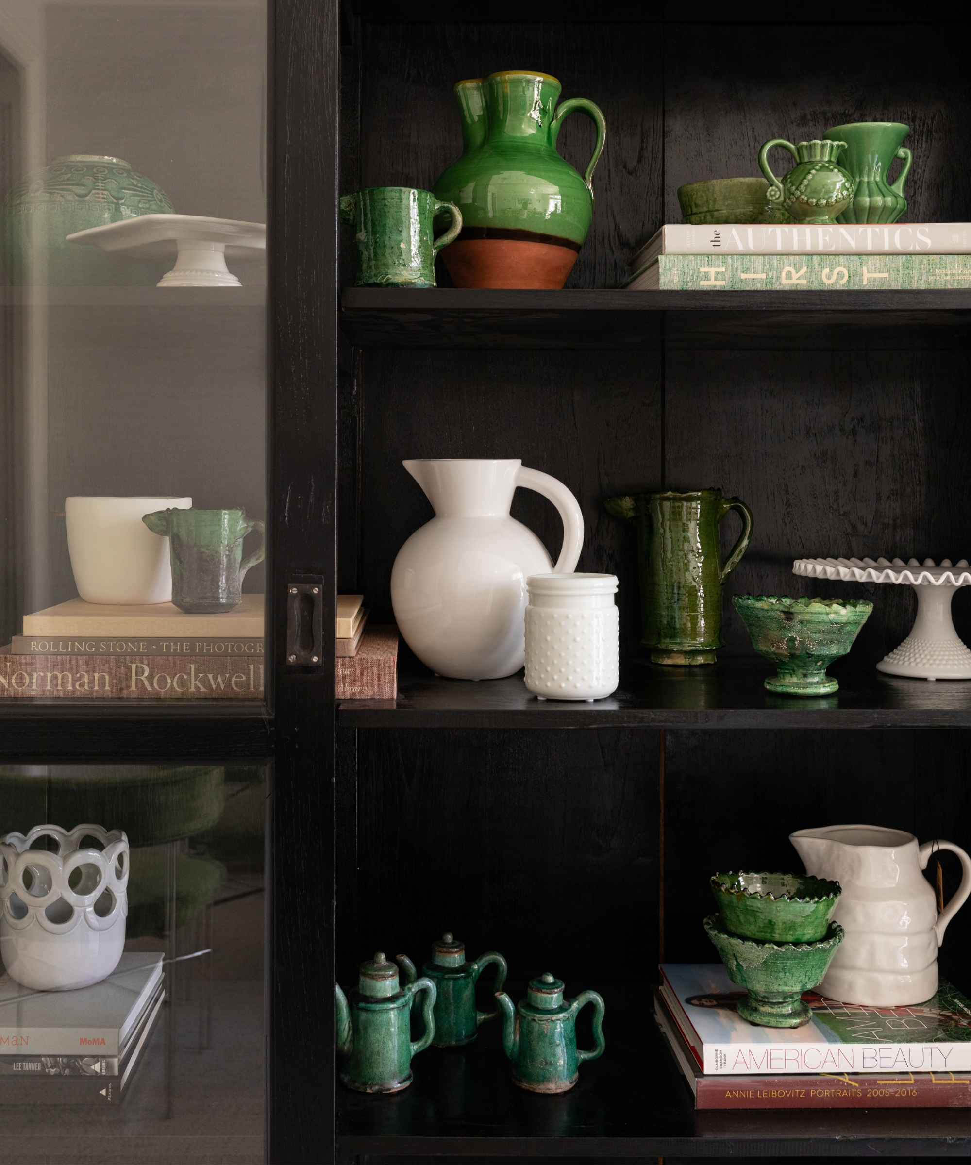 Black display cabinet styled with layered green ceramics, white pottery, books, and glass doors in a neutral interior.