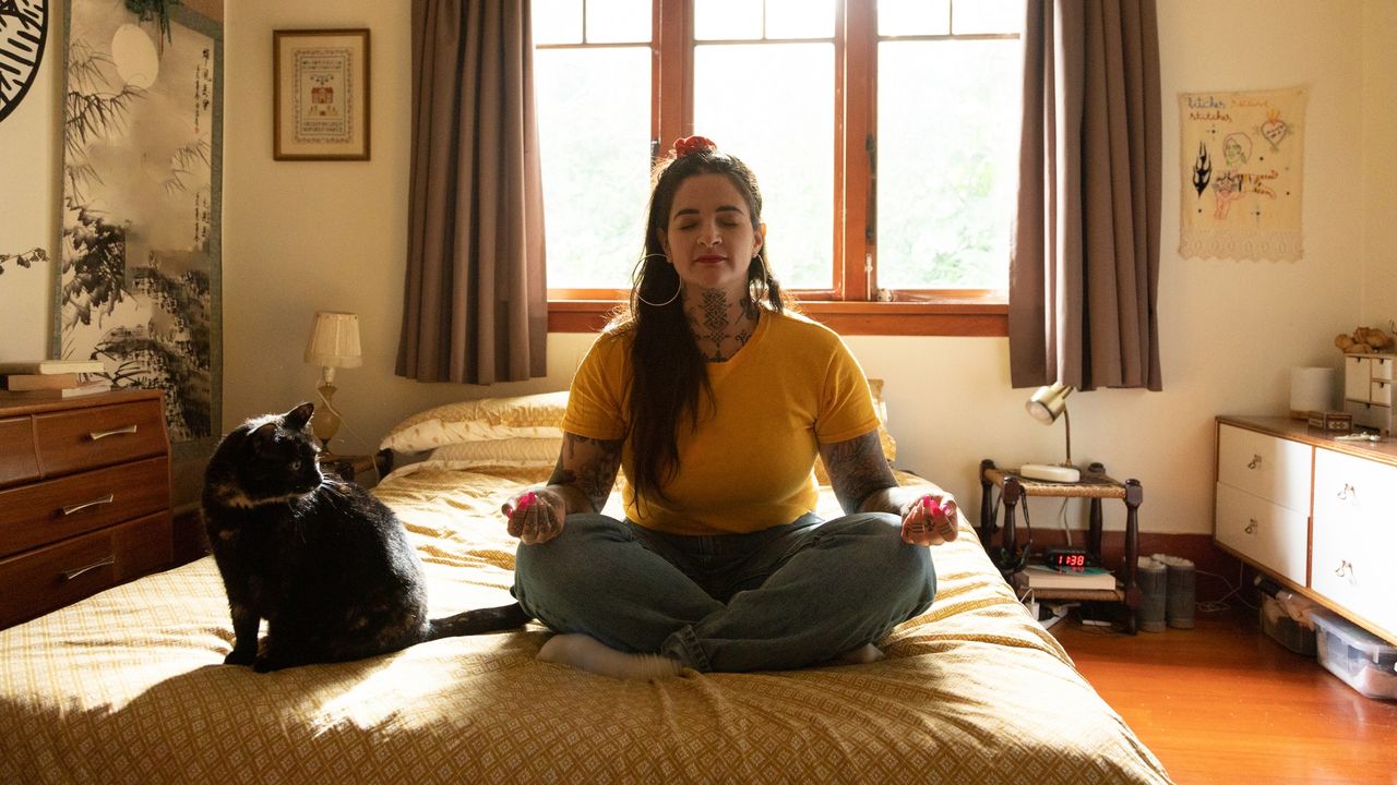 A woman in a yellow shirt sits on her bed in a meditative pose. Beside her sits a black cat.