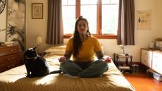 A woman in a yellow shirt sits on her bed in a meditative pose. Beside her sits a black cat.