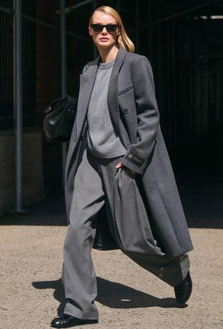 One of the best jacket coat brands, Aritzia, is shown in an image of a model wearing a long gray coat over a gray crewneck sweater with gray trousers and black loafers