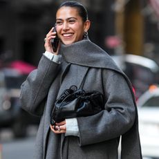 Woman wears a long grey coat with a matching scarf, black leather clutch, knee-high boots, and a denim skirt. 