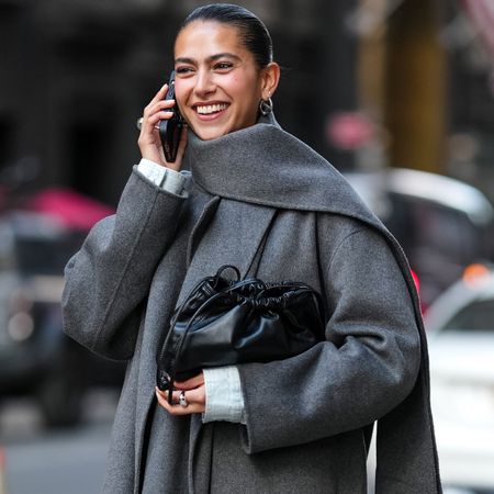 Woman wears a long grey coat with a matching scarf, black leather clutch, knee-high boots, and a denim skirt.
