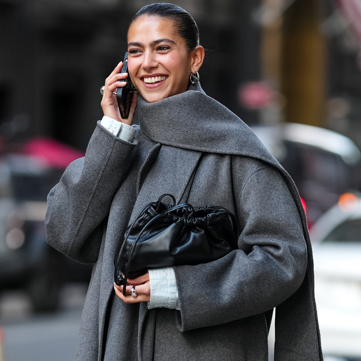 Woman wears a long grey coat with a matching scarf, black leather clutch, knee-high boots, and a denim skirt.