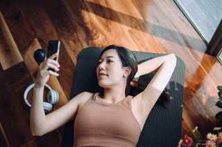 A woman using her phone while lying on a yoga mat at the gym.