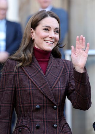 A waist up photo of Kate Middleton wearing a burgundy plaid coat and waving and smiling