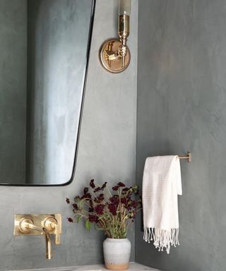 Moody bathroom detail with wall mirror, ceramic vase of flowers, and brass hardware.