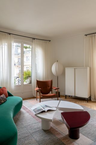 Image of a modern, white living room. There is a silk lantern floor lamp, a brown accent chair, and a white chest in the back corner by the window. In the center of the room, there is a curved couch in a green jewel tone. In front of the couch is a white and magenta pinwheel table.