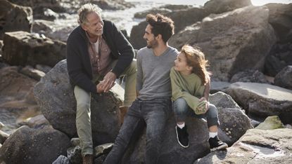 A grandfather, son and granddaughter smile and talk while sitting on a rock at oceanside.