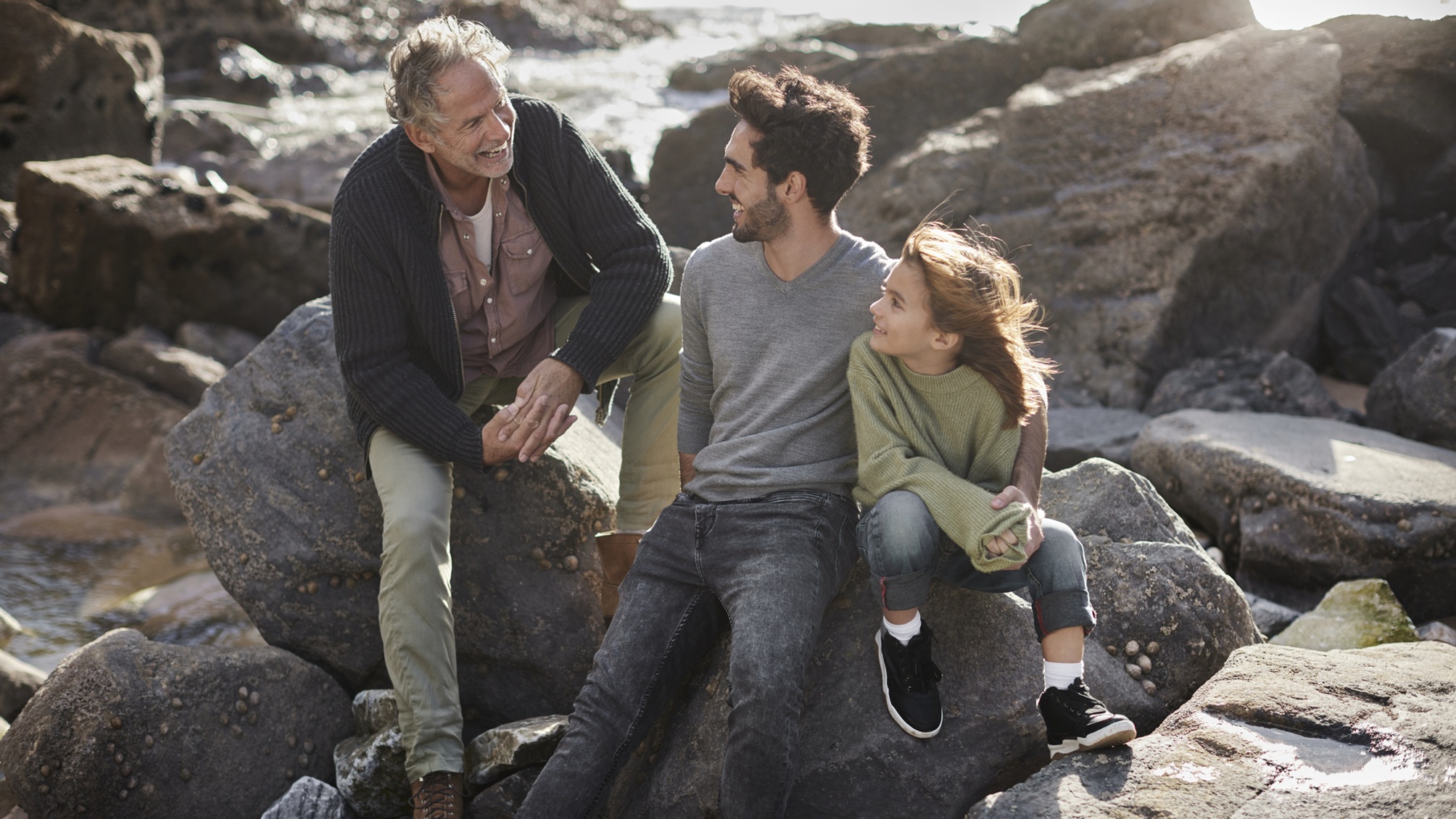 A grandfather, son and granddaughter smile and talk while sitting on a rock at oceanside.