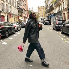 Raissa Tchoulague on the street in Paris wearing a chic fall outfit, styled with dark blue denim jeans, a longline black leather jacket, loafers, a pink and red handbag, and a black headscarf.