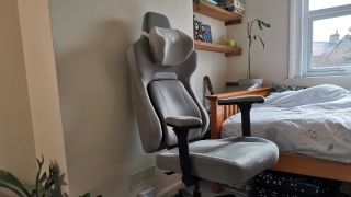 The ThundercoreX3 Core gaming chair in grey in front of a pale wall, next to a wooden bed frame