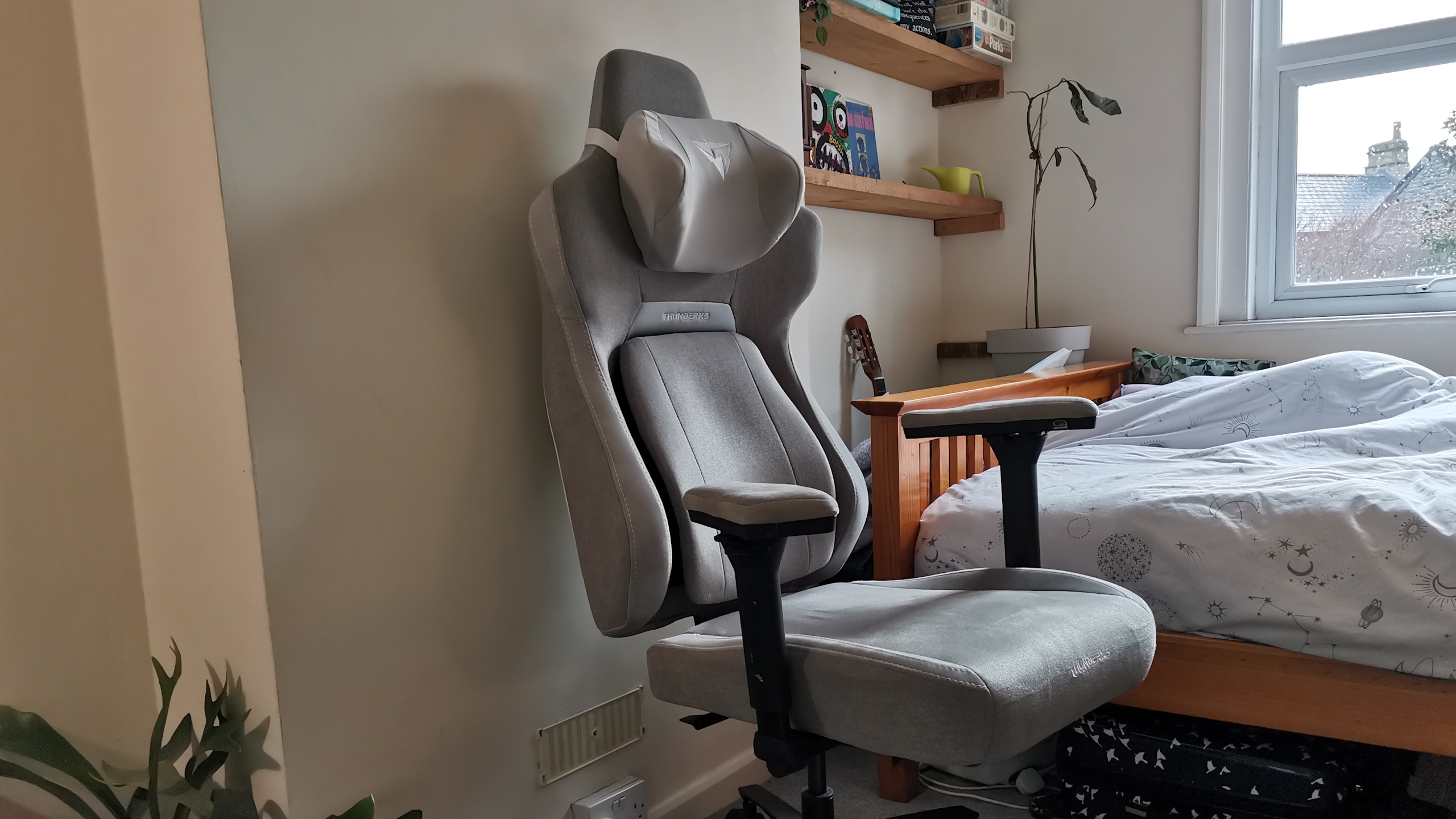 The ThundercoreX3 Core gaming chair in grey in front of a pale wall, next to a wooden bed frame