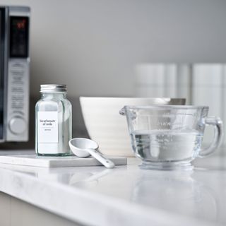 jug of water, baking soda, spoon on a worktop in front of a microwave