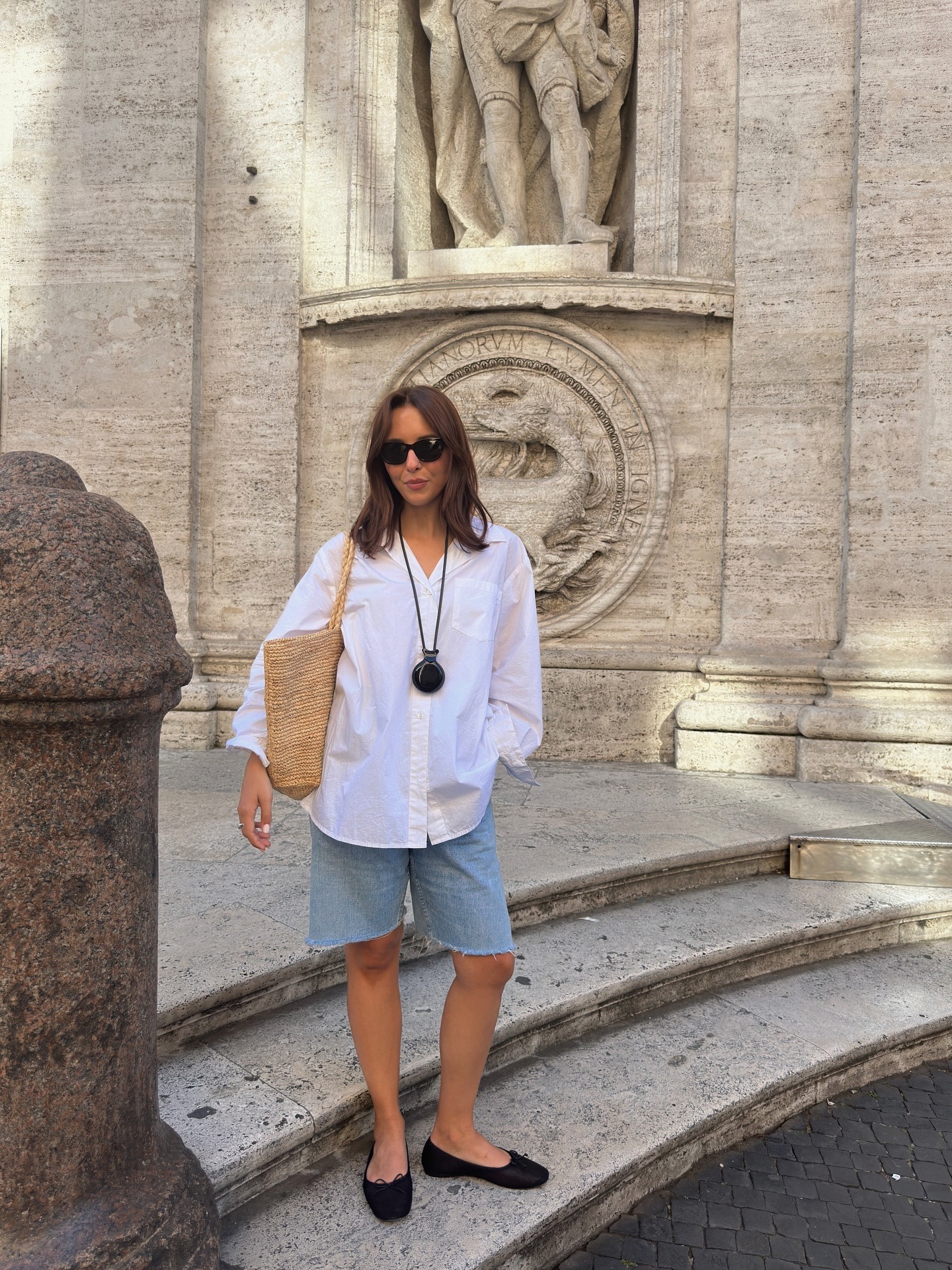 Fashion person and stylish Debora Brosa wears a white shirt, classic denim, a straw bag and a rope necklace. She is stood on steps looking at the camera in a recent trip away on a recent image on her Instagram