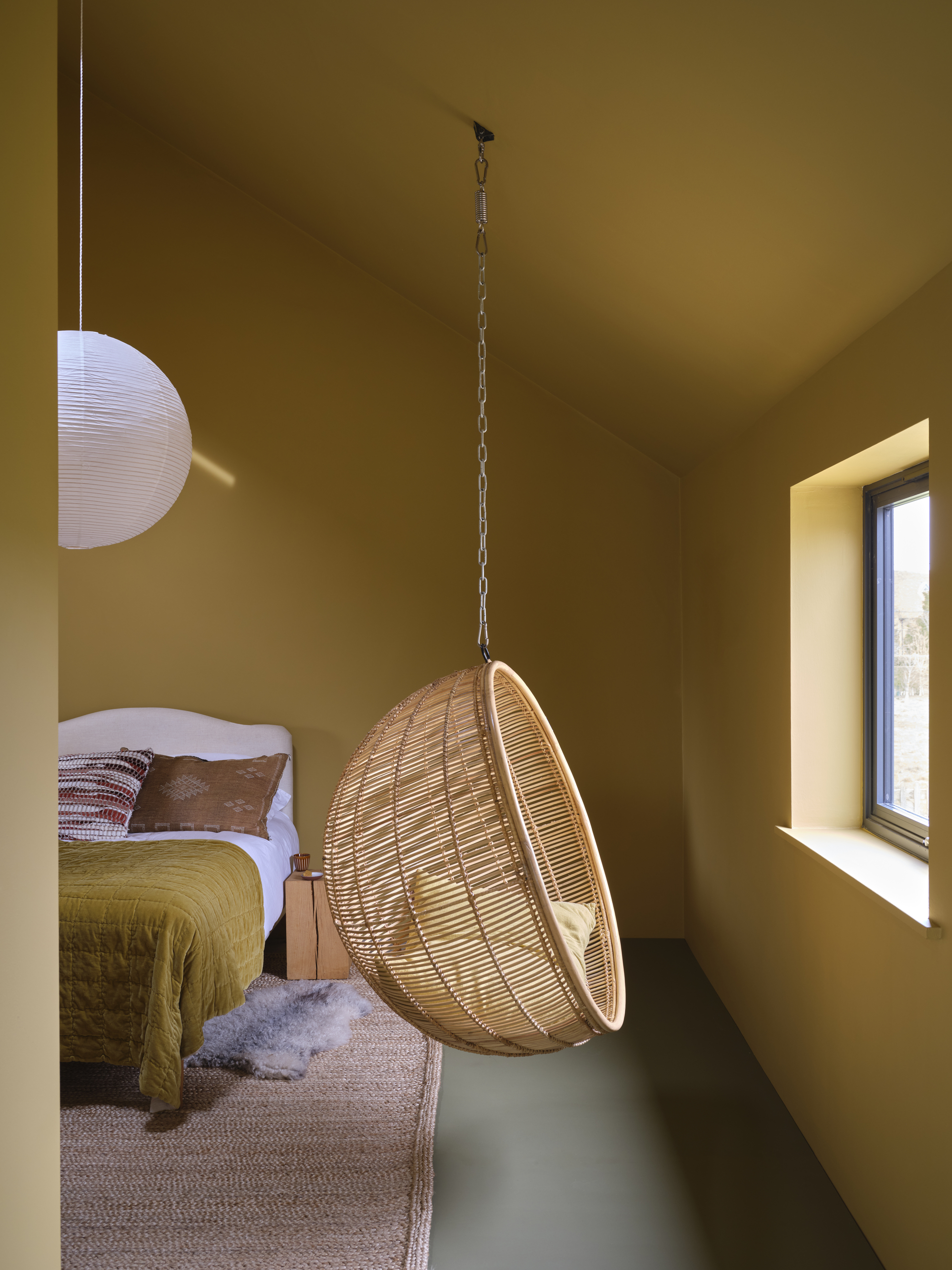 Image of a yellow bedroom with an A-frame ceiling. There is a small bed with yellow bedding and a white paper lantern hanging over it. There is also a wicker swing seat facing the window in the room.
