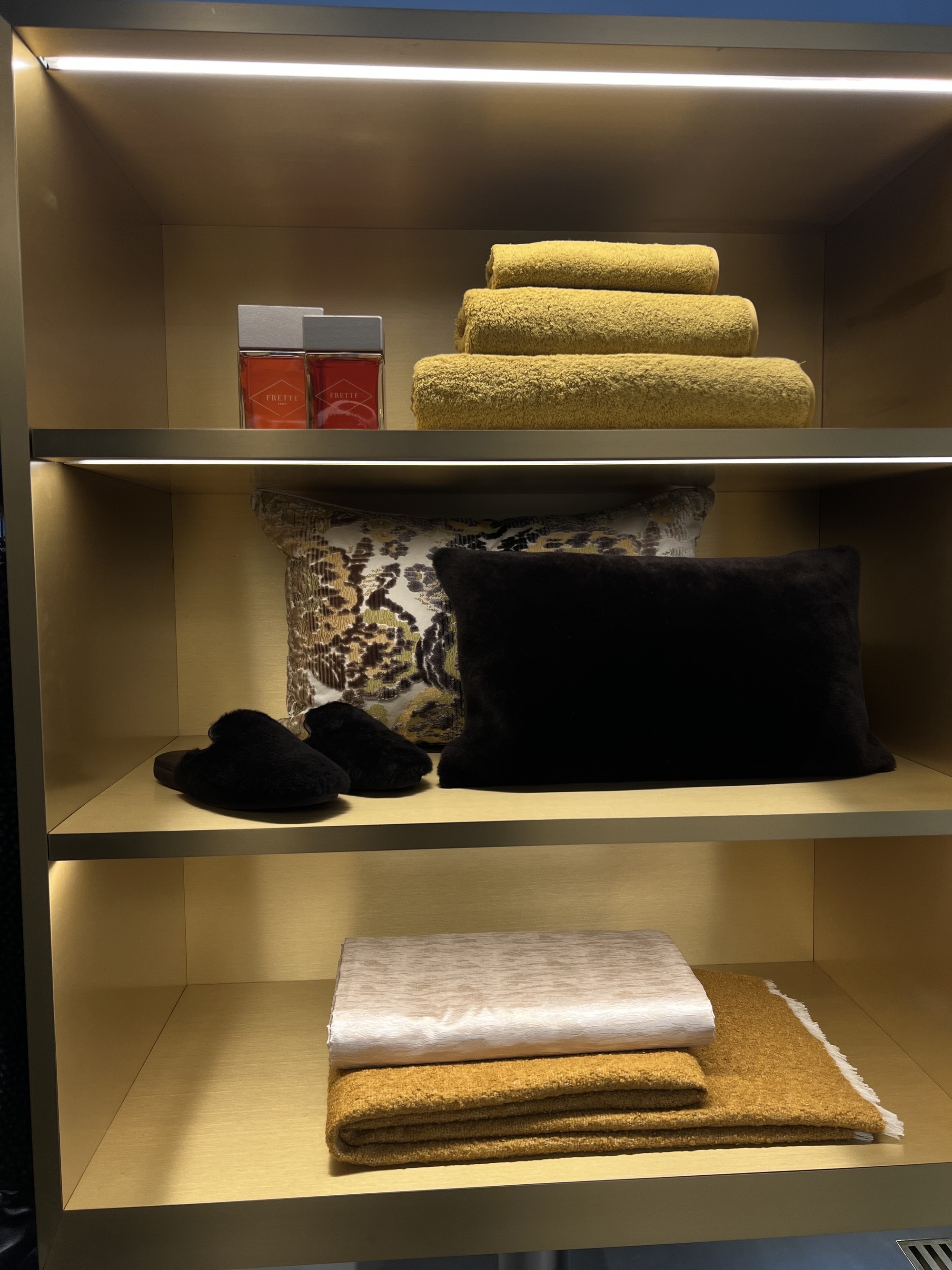 Image of luxury bedding and towel (in shades of orange, yellow, black, and light pink) displayed on a golden shelf.