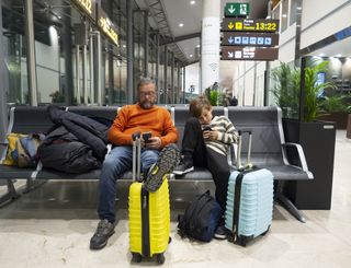 Father and tween son sitting in an airport lounge, next to wheelie suitcases, waiting for a flight