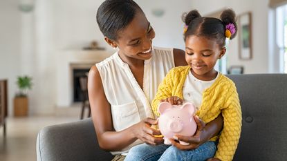 A smiling mom holds her smiling young daughter and a piggy bank on her lap.