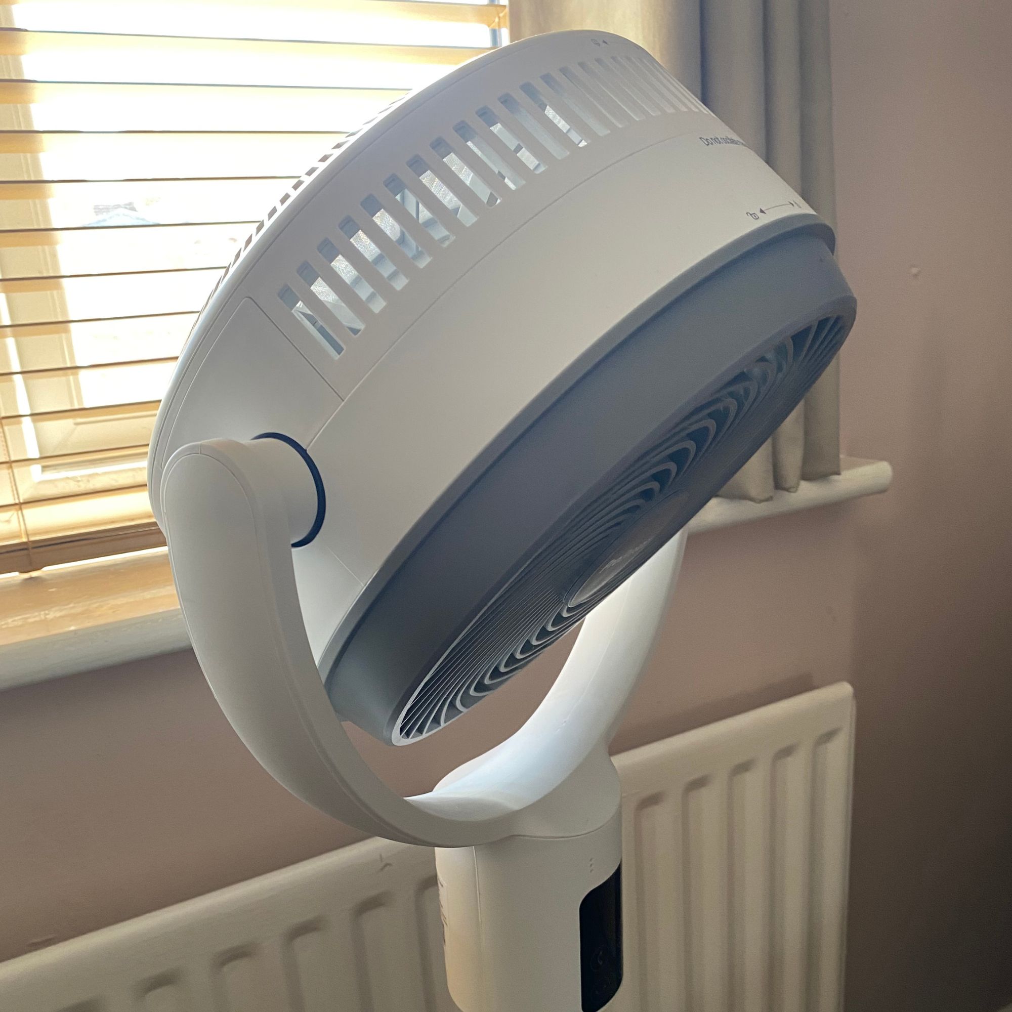 Pro Breeze Airflo 43'' Hybrid Pedestal Fan in the maximum downward vertical oscillation position in a bedroom with plaster pink walls, radiator and wooden blinds